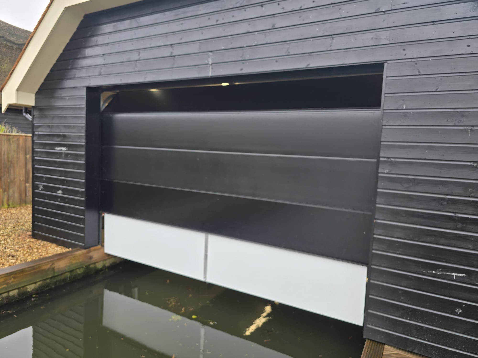 Bespoke sectional garage door installed by OGD on a dark timber-clad boathouse